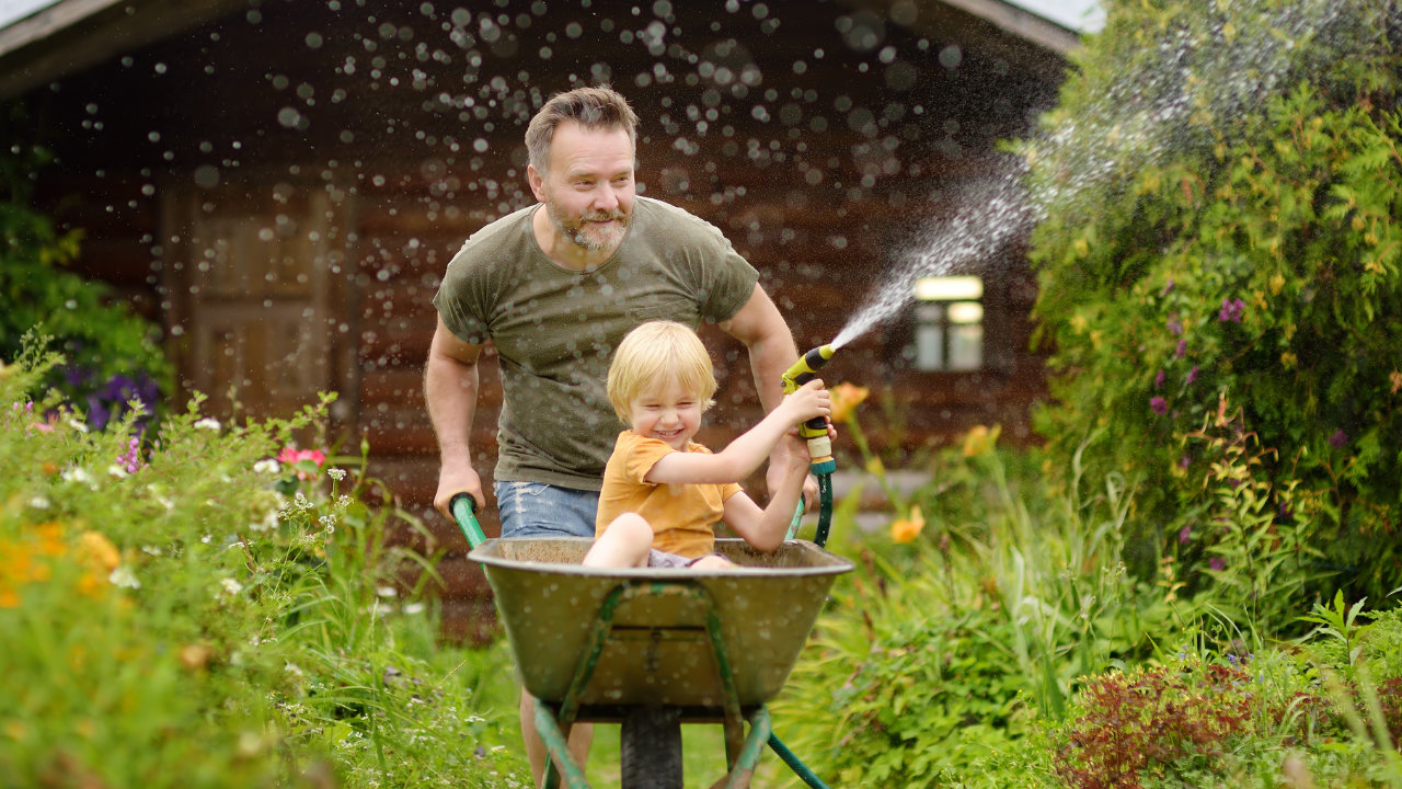 Père pousse un enfant dans une brouette dans le jardin, l’enfant arrose avec un tuyau