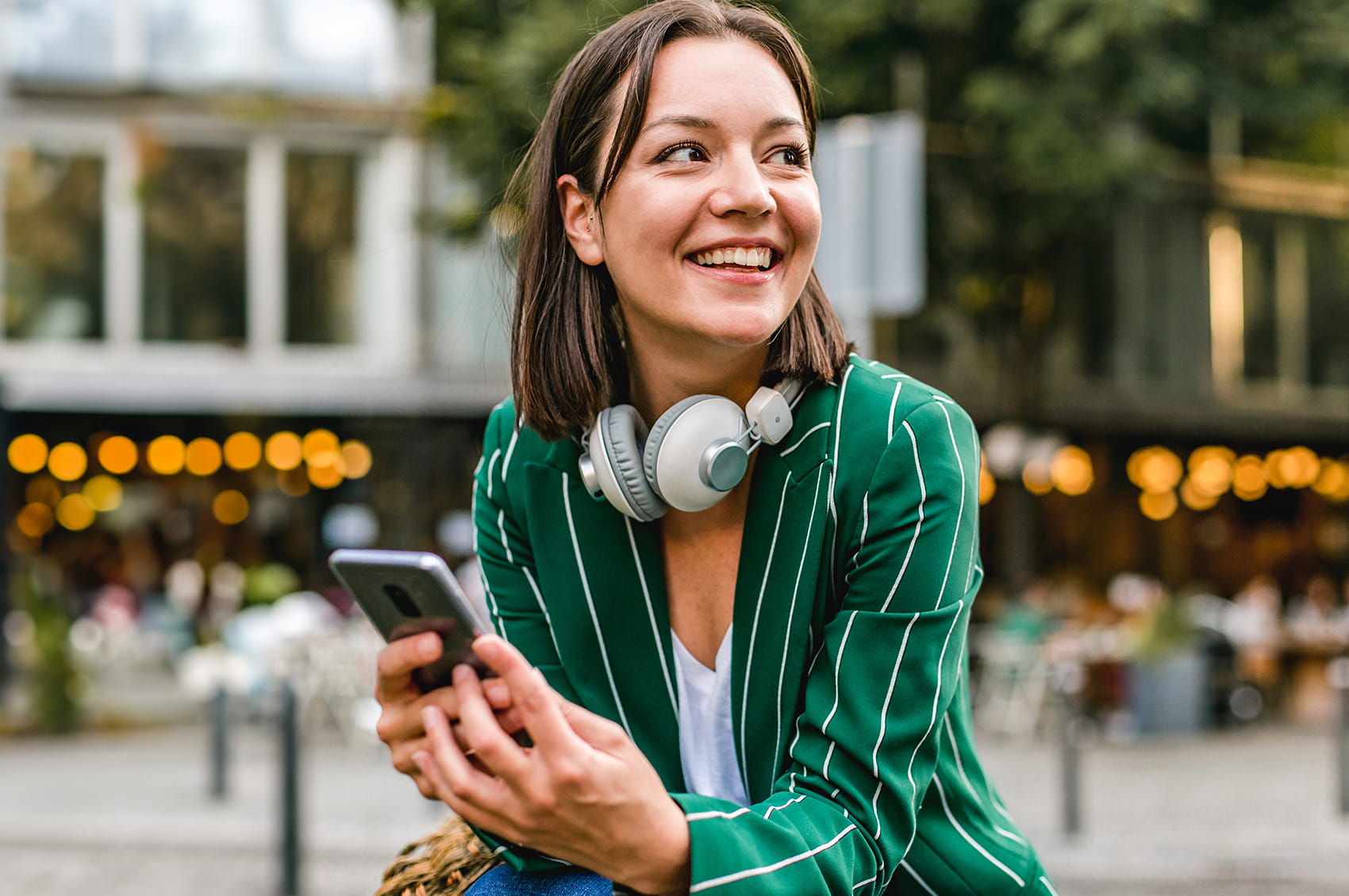 Femme assise à l’extérieur tient un smartphone et porte un casque autour du cou