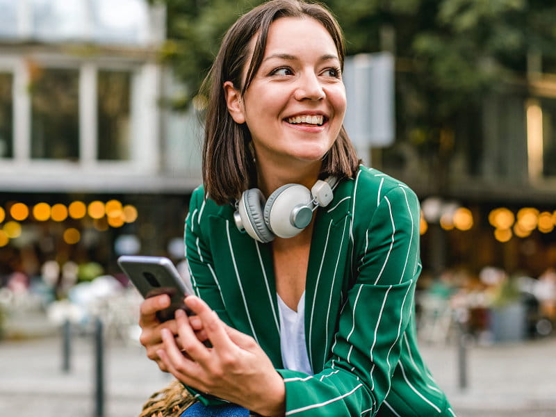 Femme assise à l’extérieur tient un smartphone et porte un casque autour du cou