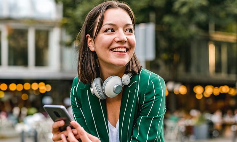 Femme assise à l’extérieur tient un smartphone et porte un casque autour du cou
