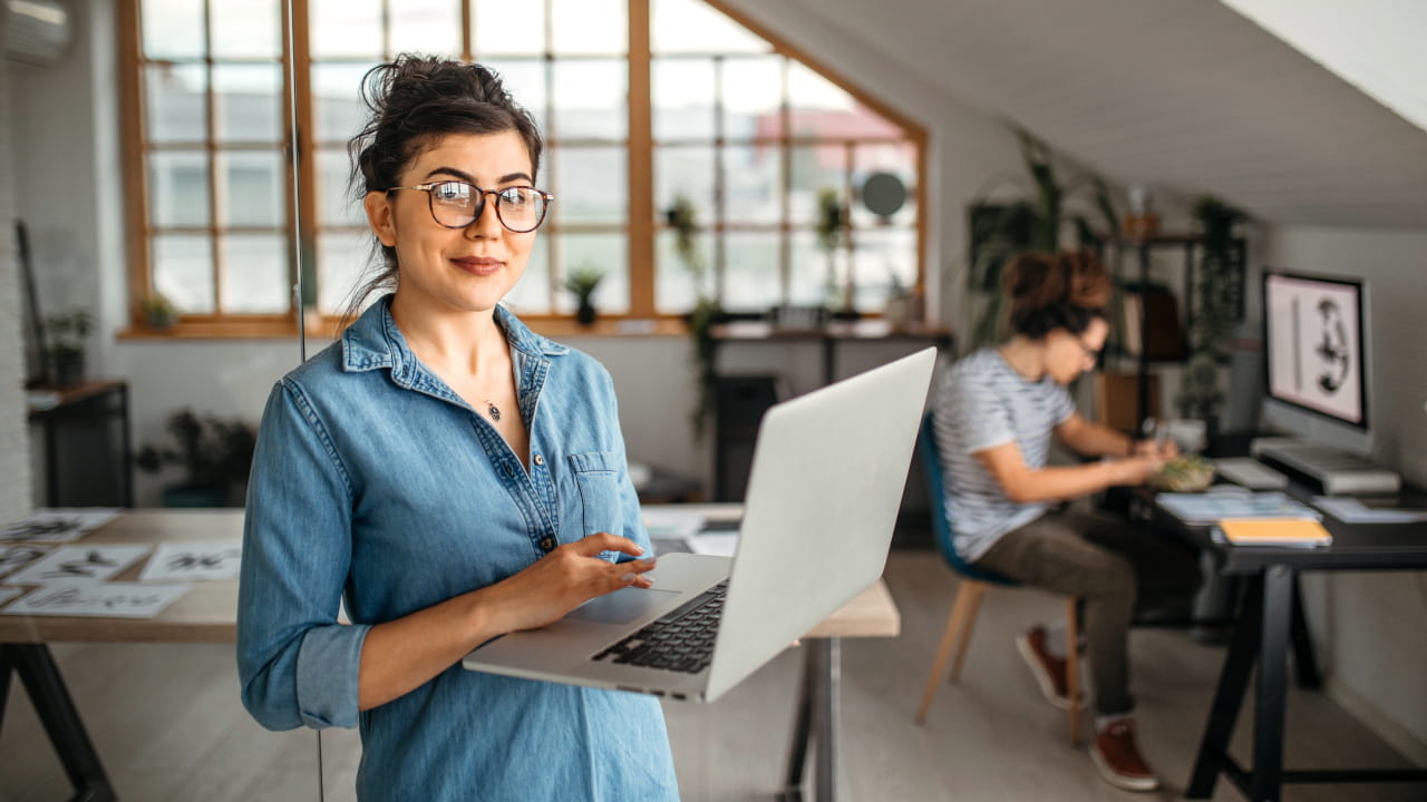 Une femme travaille sur un ordinateur portable dans un bureau; une autre personne travaille à un bureau en arrière-plan.