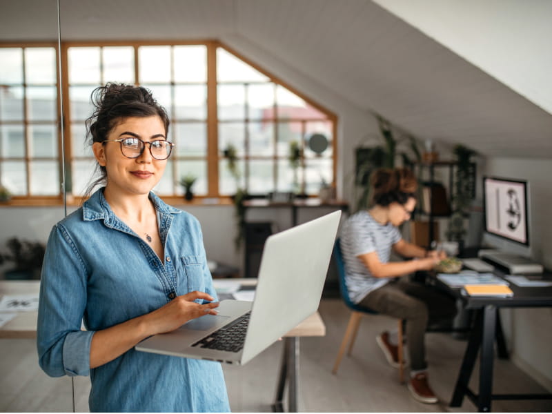 Une femme travaille sur un ordinateur portable dans un bureau; une autre personne travaille à un bureau en arrière-plan.