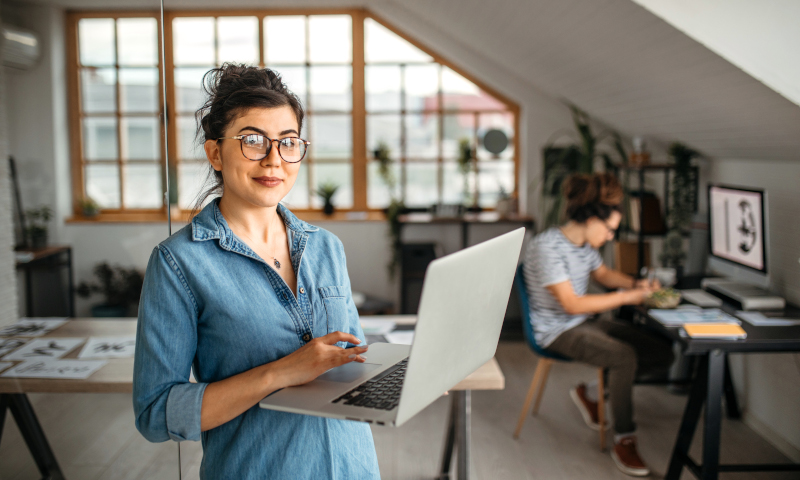 Une femme travaille sur un ordinateur portable dans un bureau; une autre personne travaille à un bureau en arrière-plan.