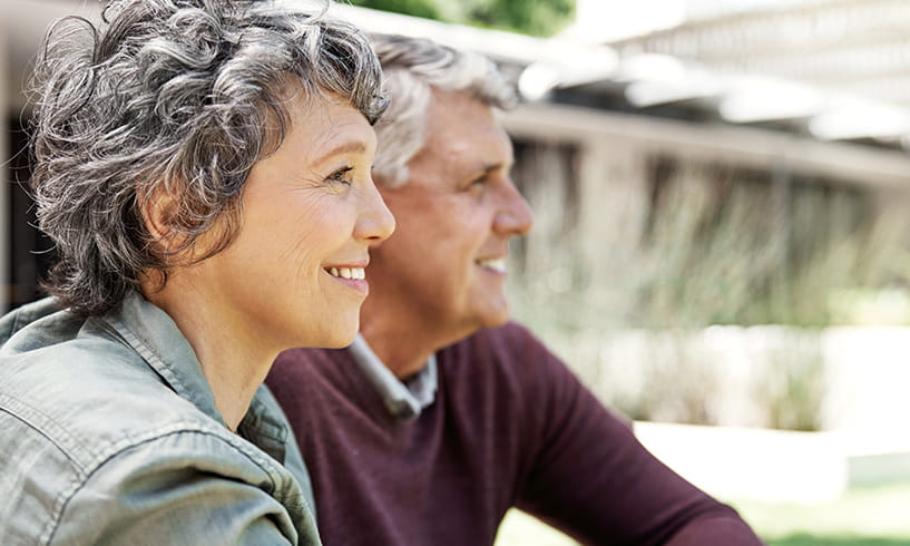 Un couple âgé souriant est assis dehors et regarde ensemble au loin.