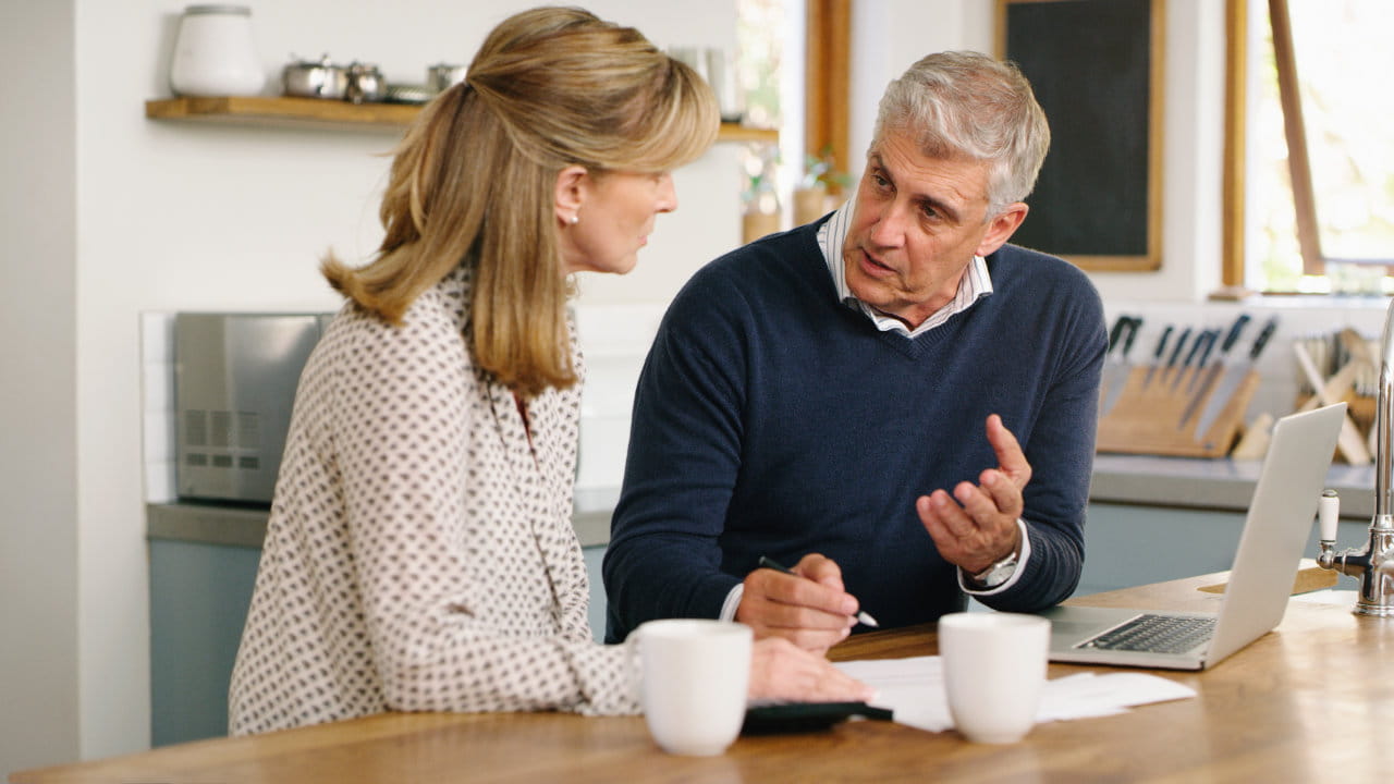 Deux adultes discutent de documents à une table de cuisine. Un ordinateur portable et deux tasses sont sur la table.