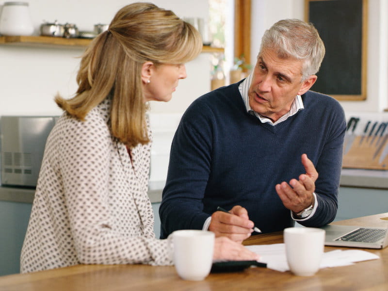 Deux adultes discutent de documents à une table de cuisine. Un ordinateur portable et deux tasses sont sur la table.