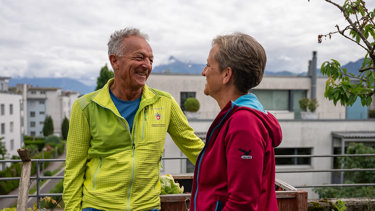 Des personnes en préretraite discutent en souriant à l’extérieur devant un quartier résidentiel avec un paysage de montagnes.