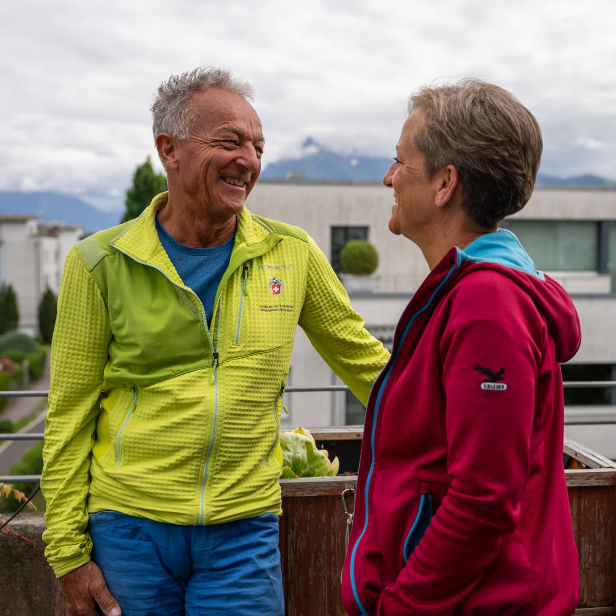 Des personnes en préretraite discutent en souriant à l’extérieur devant un quartier résidentiel avec un paysage de montagnes.