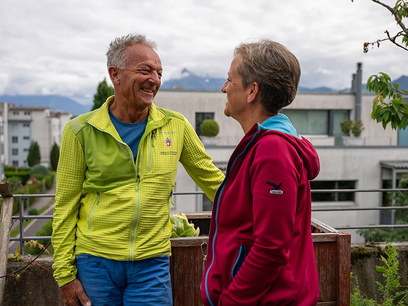 Des personnes en préretraite discutent en souriant à l’extérieur devant un quartier résidentiel avec un paysage de montagnes.
