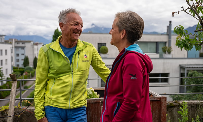 Des personnes en préretraite discutent en souriant à l’extérieur devant un quartier résidentiel avec un paysage de montagnes.